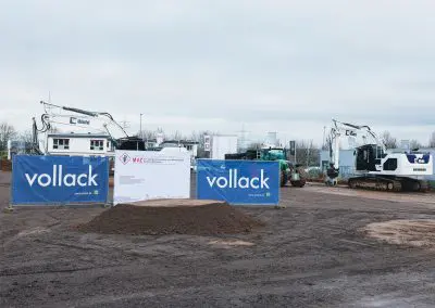 Bild des Erdhaufens vom Spatenstich ohne Personen. Im Hintergrund sind Bauzäune mit Werbebannern des MVZ und der Firma Vollack zu sehen. Weiße Bagger stehen ebenfalls im Bild.