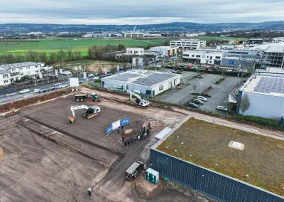 Drohnenaufnahme des Spatenstichs mit etwa 30 Personen, die sich um einen großen Erdhaufen versammelt haben. Die Spaten stecken in der Erde, während Dr. Walscheid, Geschäftsführer des MVZ Labor Koblenz, eine Rede hält. Die Teilnehmer tragen weiße Helme und es herrscht eine festliche Atmosphäre. Die Aufnahme zeigt eine größere Perspektive, sodass auch der Horizont und das umliegende Gelände sichtbar sind.