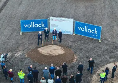 Drohnenaufnahme des Spatenstichs mit etwa 30 Personen, die sich um einen großen Erdhaufen versammelt haben. Die Spaten stecken in der Erde, während Dr. Walscheid, Geschäftsführer des MVZ Labor Koblenz, eine Rede hält. Die vier Personen hinter dem Erdhaufen tragen weiße Helme und es herrscht eine festliche Atmosphäre.