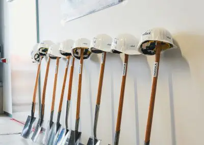 Neun Spaten lehnen an einer Wand, wobei auf jedem Spaten ein weißer Bauhelm liegt. An der Wand ist eine 3D-Ansicht des zukünftigen Neubaus zu sehen.