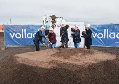 Foto vom offiziellen Spatenstich mit fünf Personen, darunter Dr. Walscheid, Geschäftsführer des MVZ Labor Koblenz. Alle tragen weiße Helme und werfen mit ihren Spaten Erde auf einen großen Erdhaufen. Sie lächeln dabei.