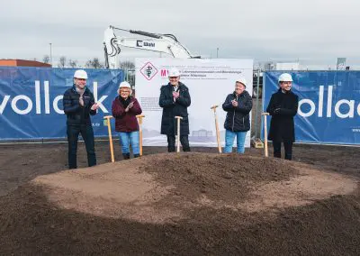Foto vom Spatenstich mit fünf Personen, darunter Dr. Walscheid, Geschäftsführer des MVZ Labor Koblenz. Alle tragen weiße Helme und stehen vor einem großen Erdhaufen. Die Spaten stecken in der Erde, während die Personen klatschen und lächeln.