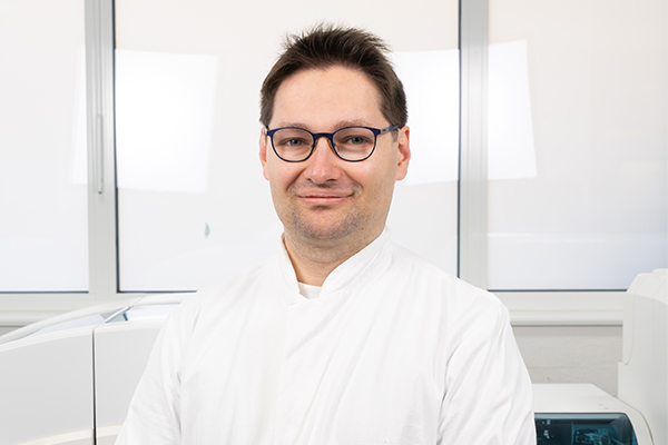 Porträtfoto von Philipp Funk, Arzt in Weiterbildung zum Facharzt für Laboratoriumsmedizin. Er blickt freundlich in die Kamera und trägt einen weißen Laborkittel. Der Hintergrund ist neutral gehalten.